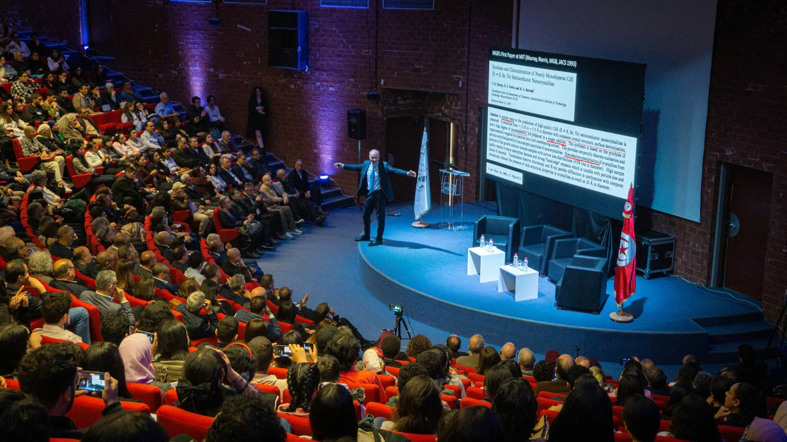 A presenter addressing a packed conference auditorium with a brightly lit stage