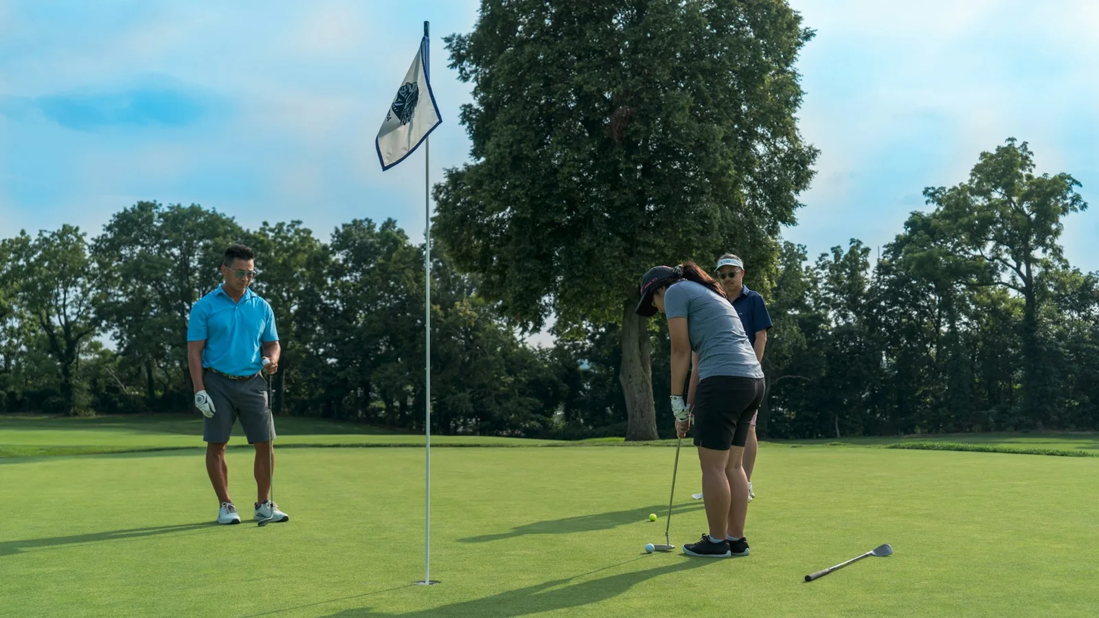 Golfers on a sunlit putting green beside a flagstick, with trees lining the course