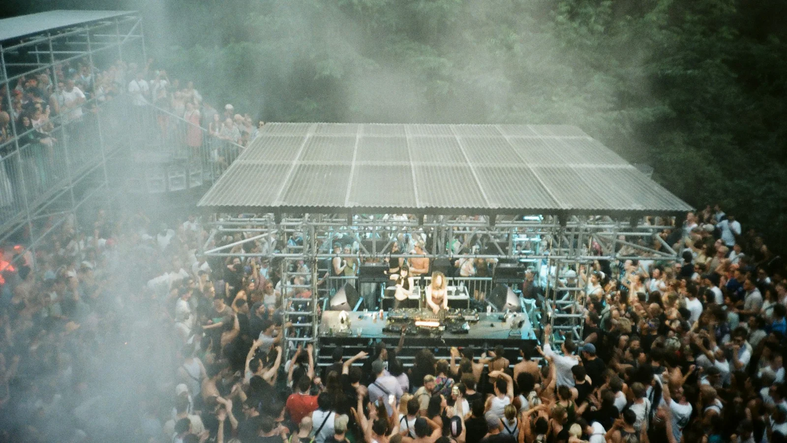A crowd gathered around a covered outdoor festival stage with truss roofing and atmospheric haze
