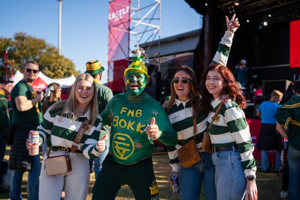Four rugby fans in green and white outfits smile and pose at an outdoor Rugby Match, with one painted green and wearing a yellow hat. A stage and other attendees are visible at Loftus Park for the Springboks vs Italy event.