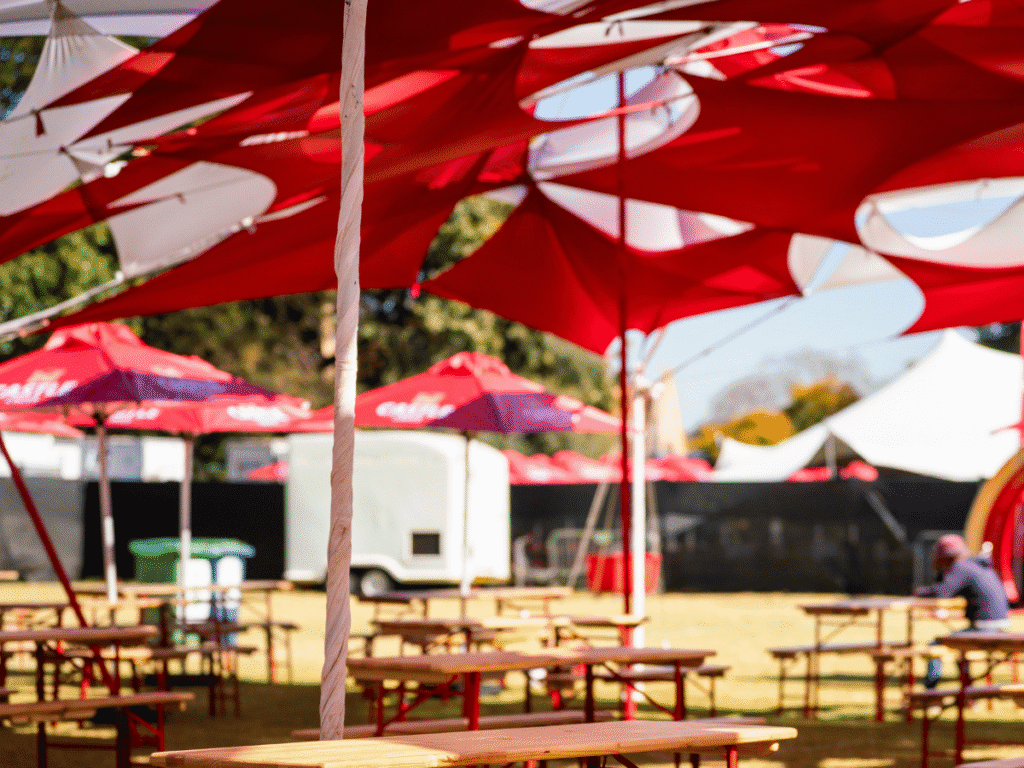 Outdoor event area at Loftus Park, featuring red and white tents and umbrellas, wooden tables and benches, with a few people relaxing in the background—perfect for enjoying the Springboks vs Italy Rugby Match.