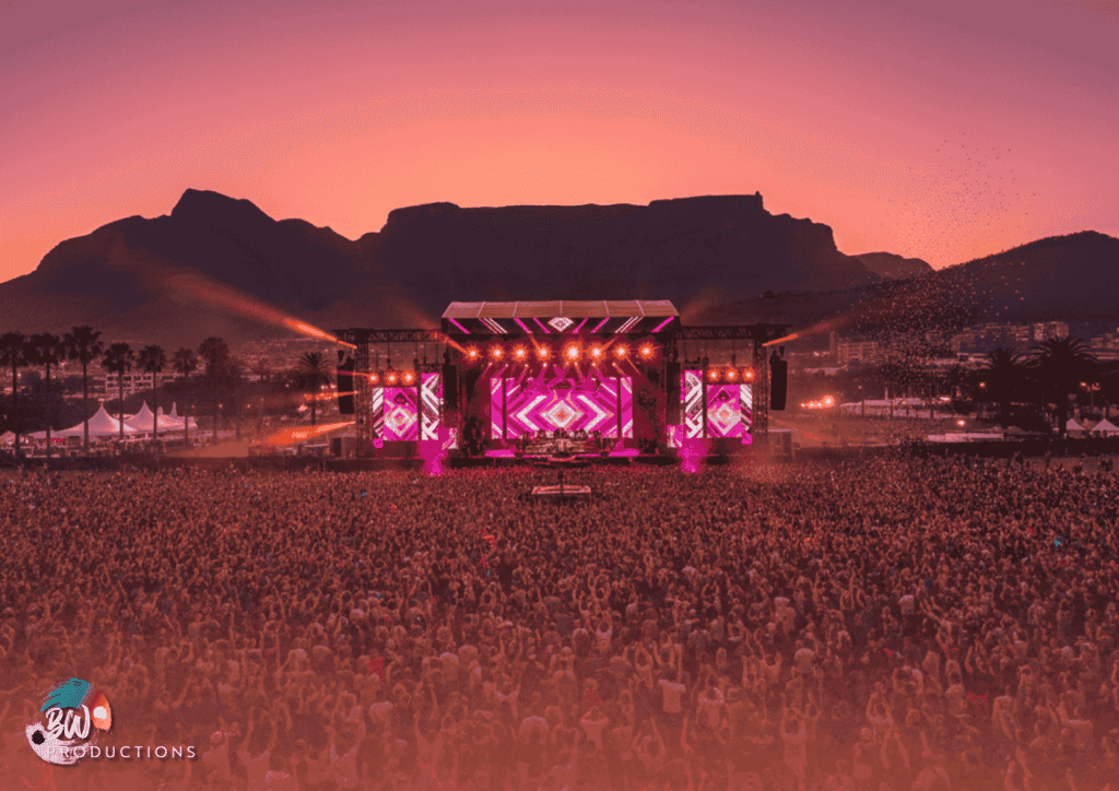Large crowd at an outdoor concert with a brightly lit stage, set against mountains at sunset.