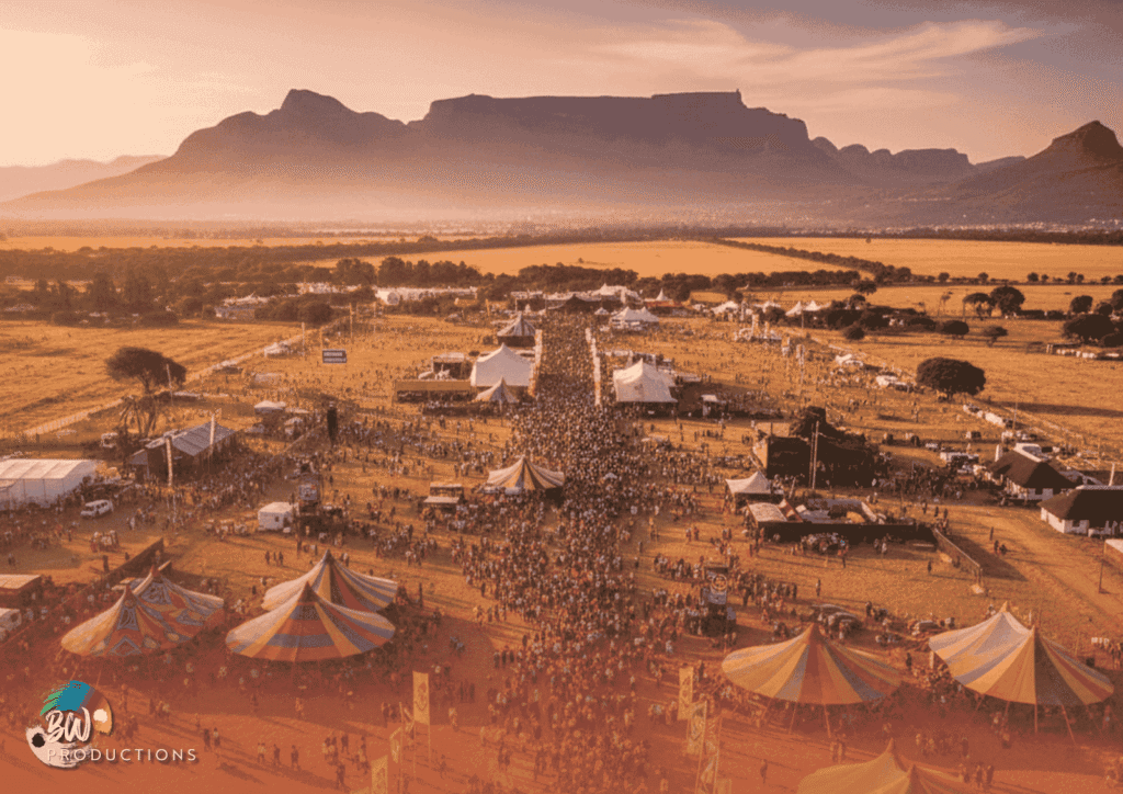 A large crowd gathers at an outdoor festival with multiple tents and stages, surrounded by fields and mountains in the background during sunset.
